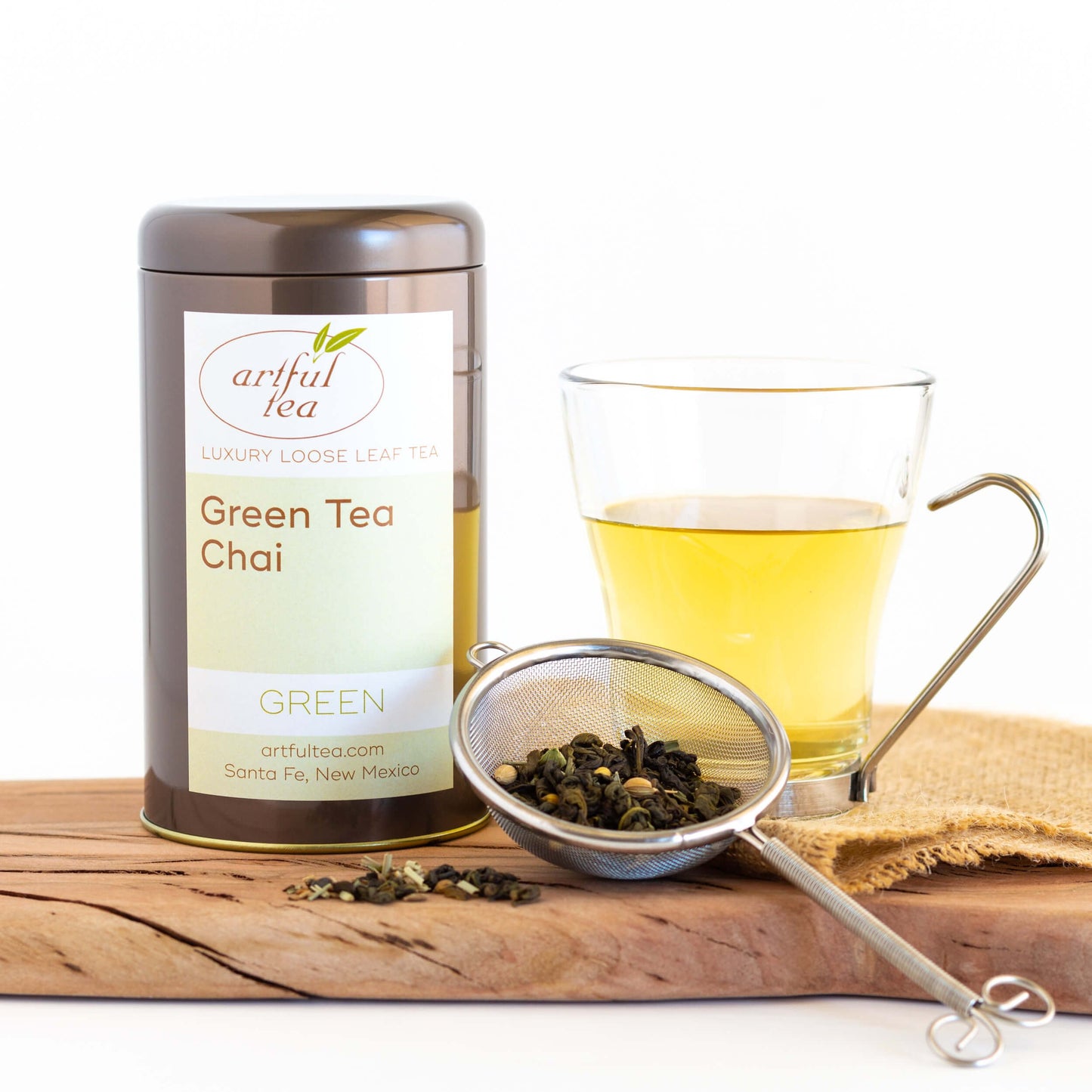 Green Tea Chai shown packaged in a brown tin next to a clear mug of brewed tea and a strainer full of loose leaf tea on top of wooden plank.