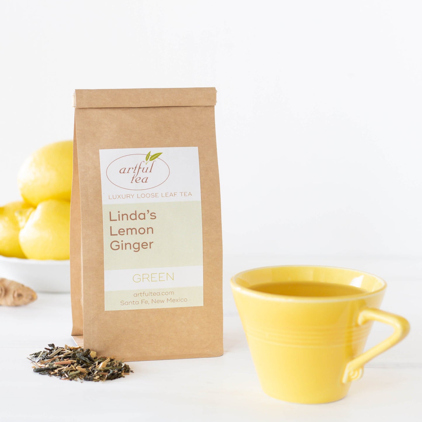 Linda's Lemon Ginger shown packaged in a brown Kraft bag beside a yellow mug full of brewed teas with a bowl of lemons in the background.