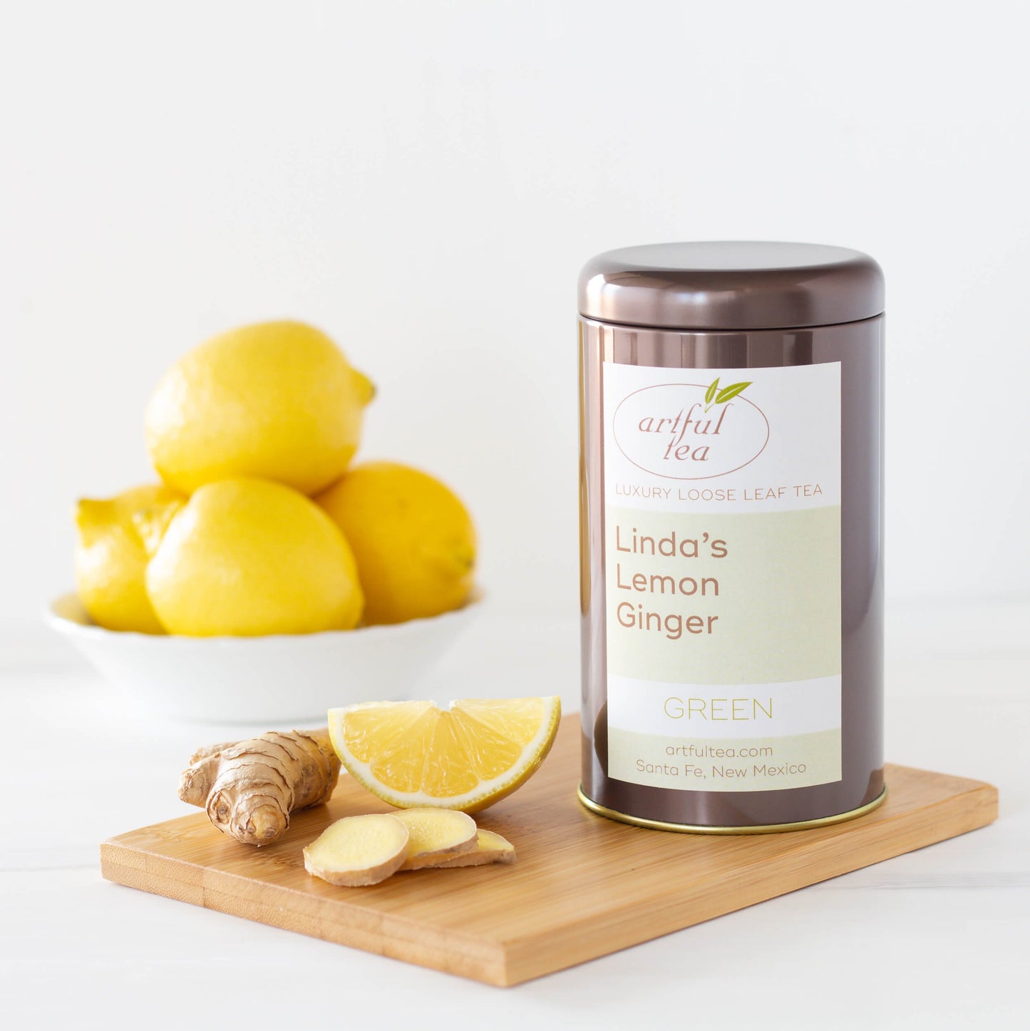 Linda's Lemon Ginger Green Tea shown packaged in a brown tin surrounded by ginger and lemon on a wooden plank.