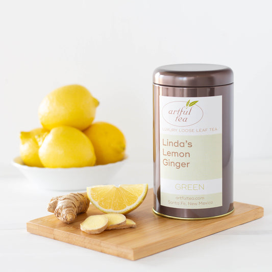 Linda's Lemon Ginger Green Tea shown packaged in a brown tin surrounded by ginger and lemon on a wooden plank.