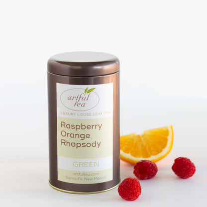 Raspberry Orange Rhapsody Green Tea shown packaged in a brown tin with a wedge of orange and three raspberries nearby