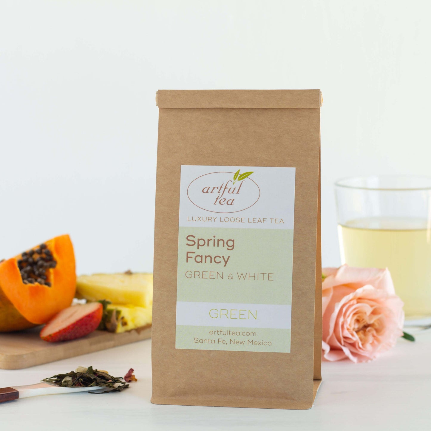 Spring Fancy Green and White Tea shown packaged in a brown Kraft bag next to a spoon of loose tea leaves, ingredients, a pink rose, and a clear cup of brewed tea.