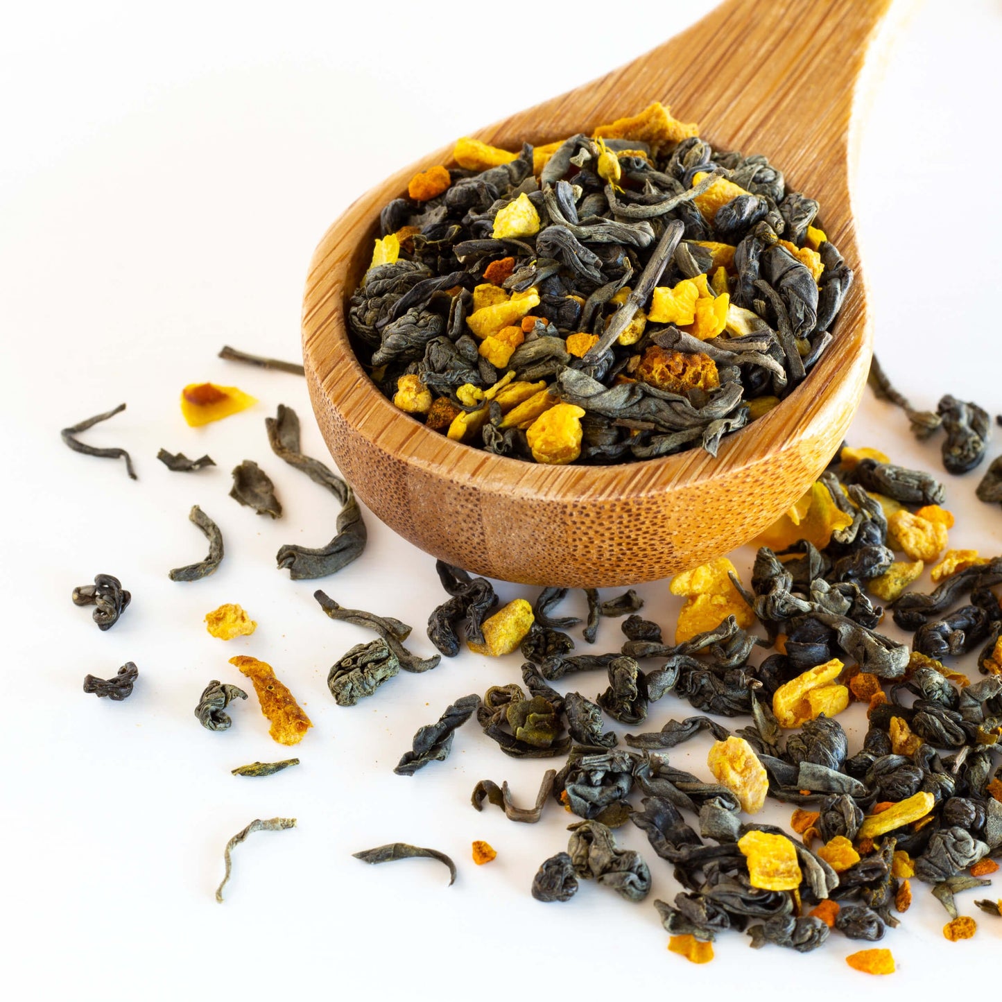 Turmeric Mango Green Tea shown as loose leaf tea in a wooden spoon with tea leaves strewn around it.