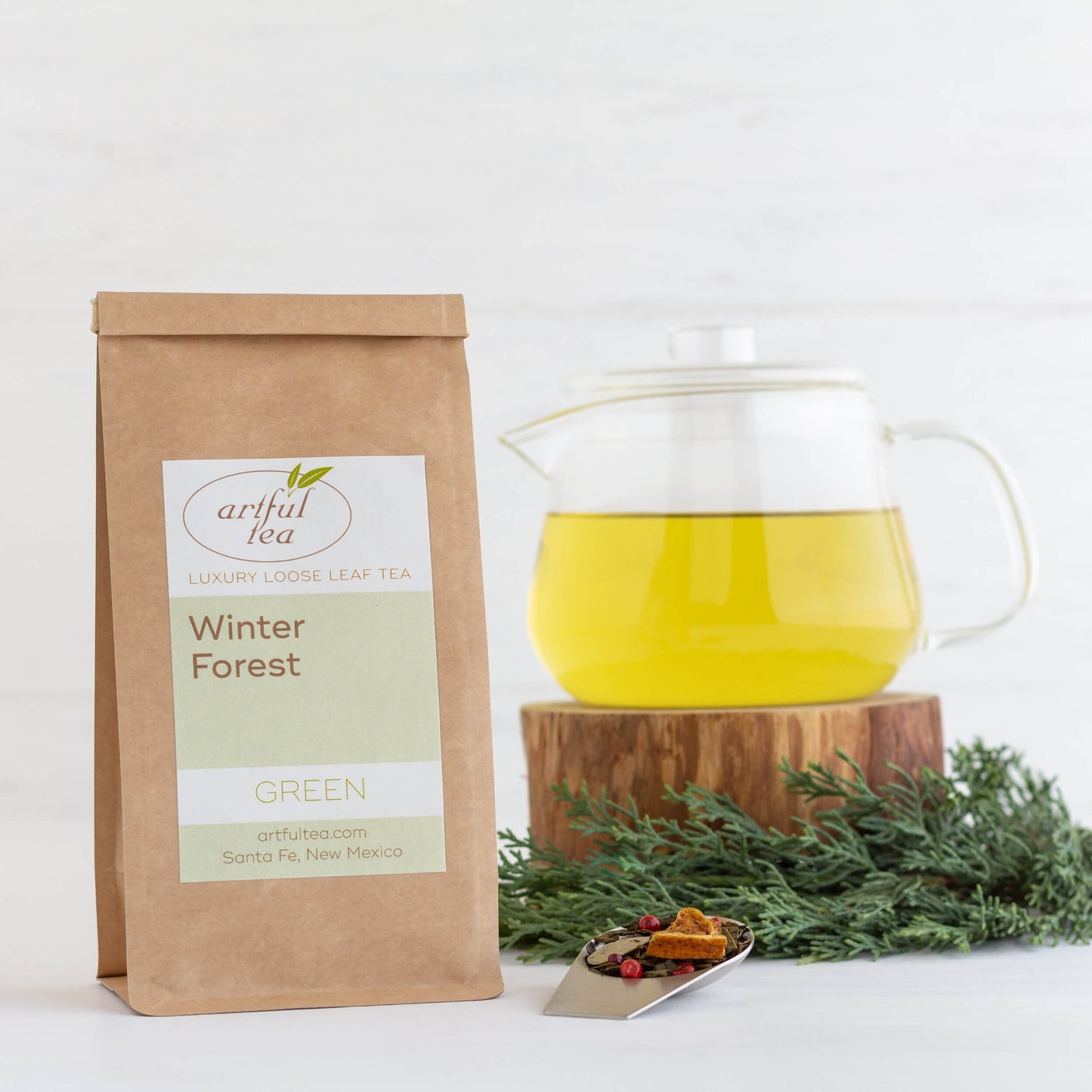 Winter Forest Green Tea shown packaged in a brown Kraft bag next to a clear teapot full of brewed tea on a piece of wood with ingredients nearby.