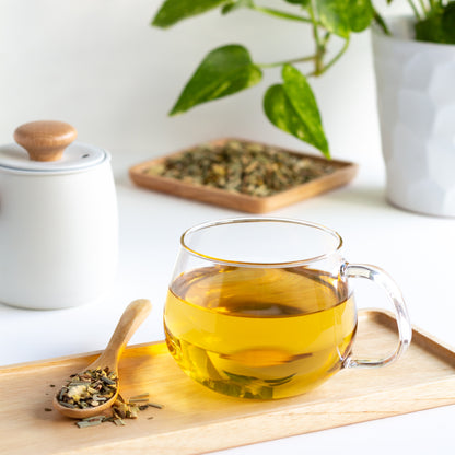 Organic Sing Your Song Herbal Tea shown brewed in a glass mug, displayed on a wooden tray. A wooden spoon with ingredients is nearby. More loose tea on a wooden dish is in the background.