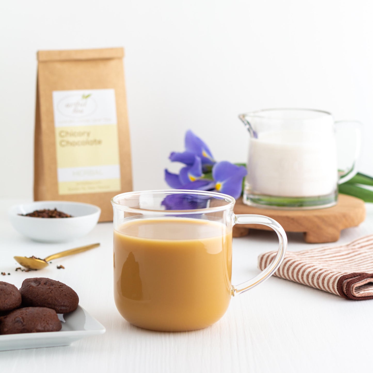 Chicory Chocolate Herbal Tea shown as a brewed tea with milk in a clear mug. Nearby is a pitcher of milk, a brown Kraft bag of Chicory Chocolate Tea, chocolate cookies, and a cloth.