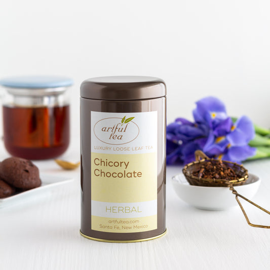 Chicory Chocolate Herbal Tea shown packaged in a brown tin. In the background is a tray of chocolate cookies, a clear mug of brewed tea, purple flowers, and a scoop of loose Chicory Chocolate Tea in a white bowl.