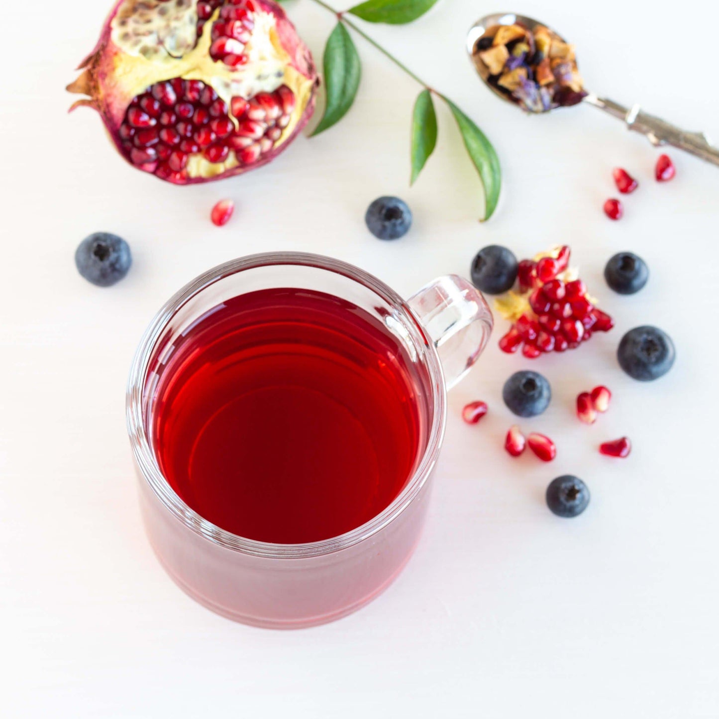 Blueberry Pomegranate Herbal Tea shown from above as brewed tea in a glass mug, surrounded by loose blueberries, an open pomegranate, and a spoon of loose tea
