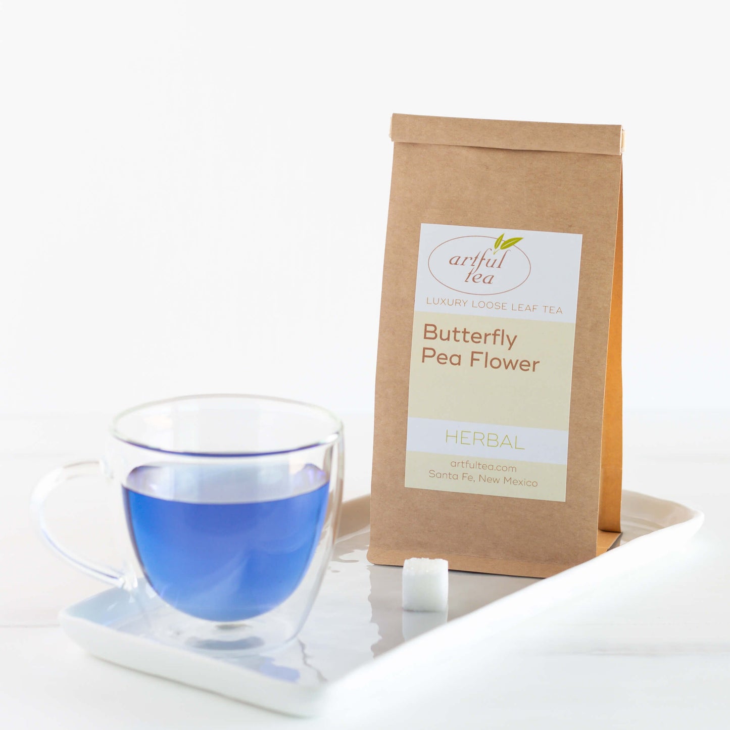 Butterfly Pea Flower Herbal Tea shown packaged in a brown Kraft bag on a white tray with a clear mug of brewed tea that is blue and a sugar cube.
