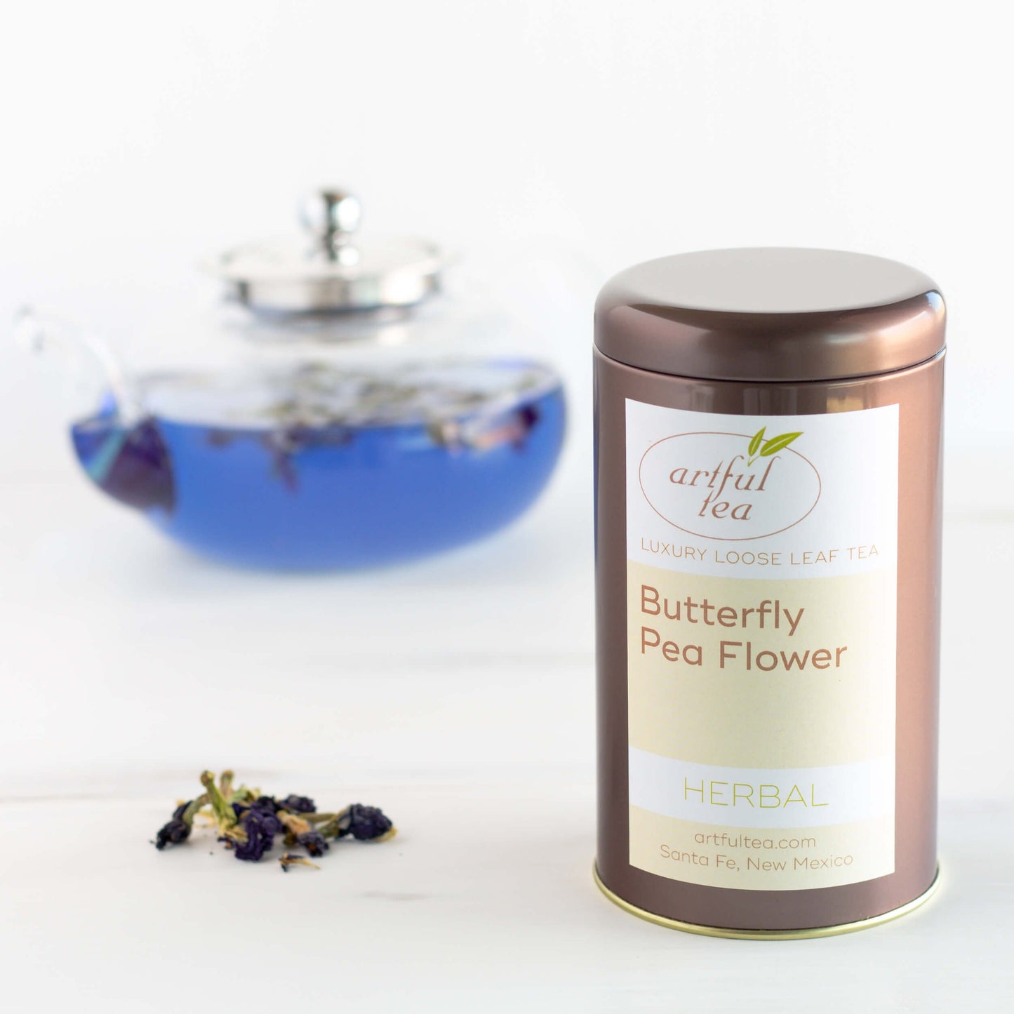 Butterfly Pea Flower Herbal Tea shown packaged in a brown tin next to a small pile of loose tea. In the background is a clear teapot with brewed tea that is blue.