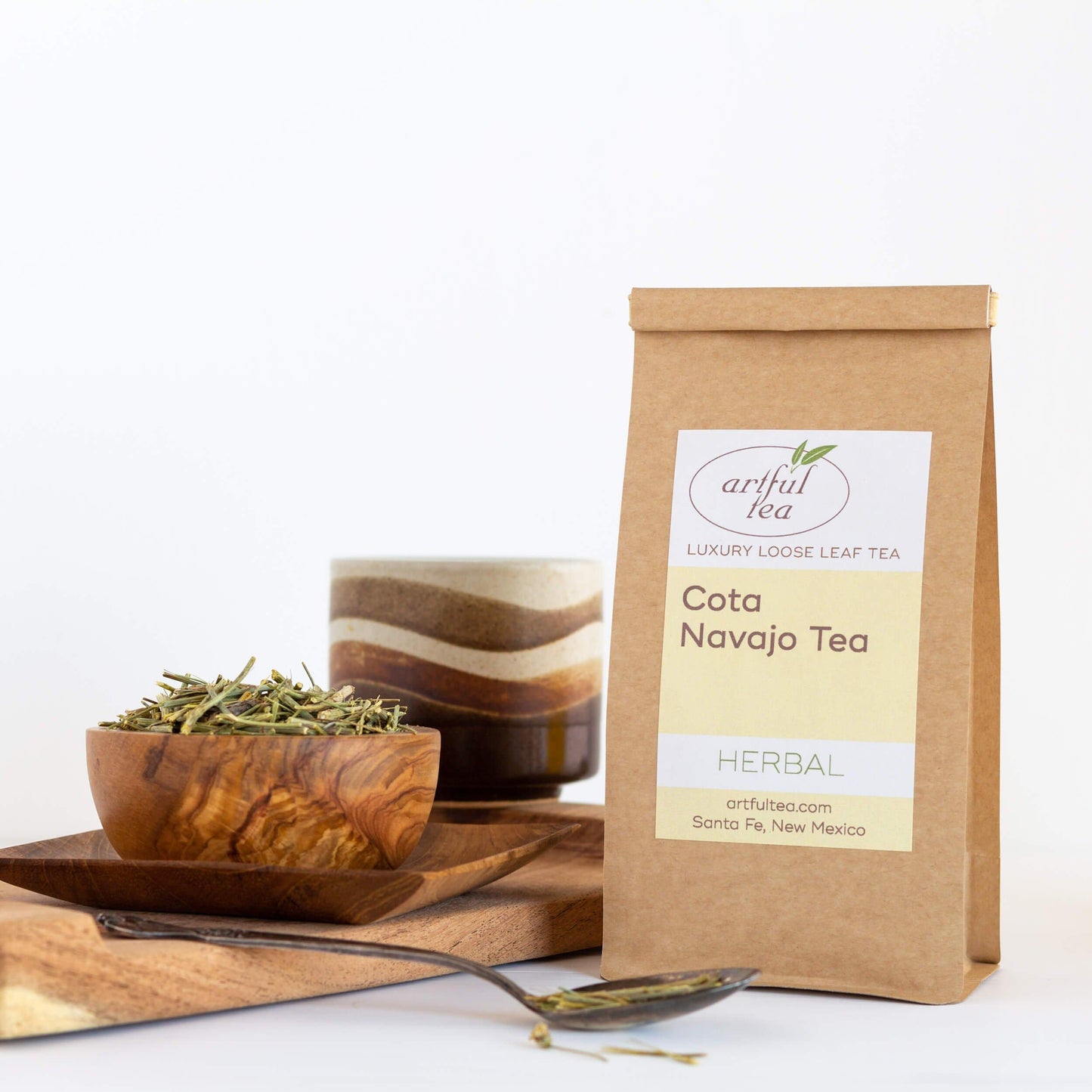 Cota Navajo Herbal Tea shown packaged in a brown Kraft bag beside a wooden bowl of loose tea on top of wooden trays, and a pottery mug in the background.