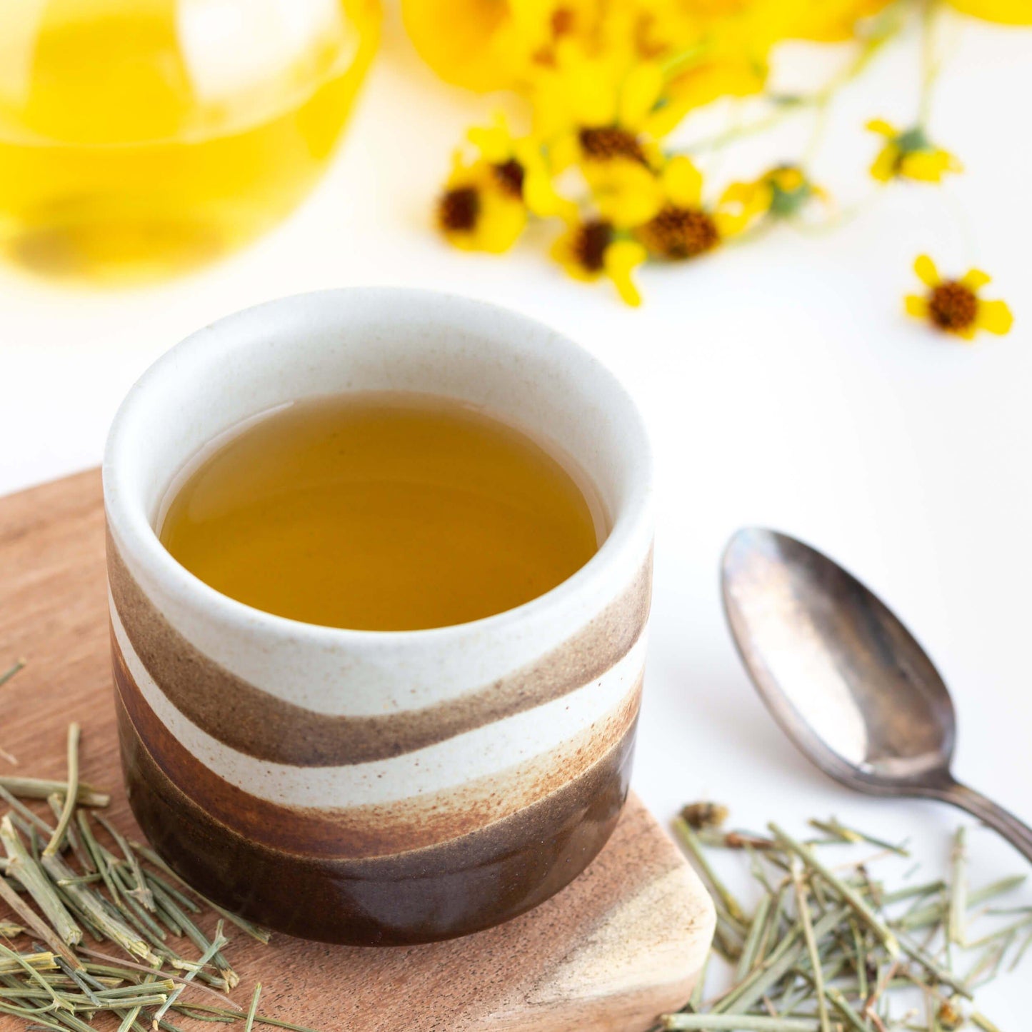 Cota Navajo Tea shown as brewed tea in a brown and white pottery cup with yellow flowers, loose grassy twigs, and a spoon nearby