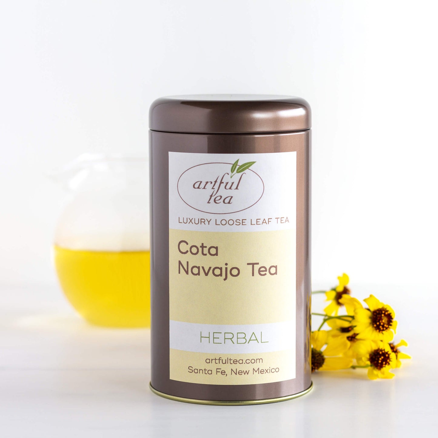 Cota Navajo Herbal Tea shown packaged in a brown tin beside a clear pitcher of brewed tea and yellow flowers.