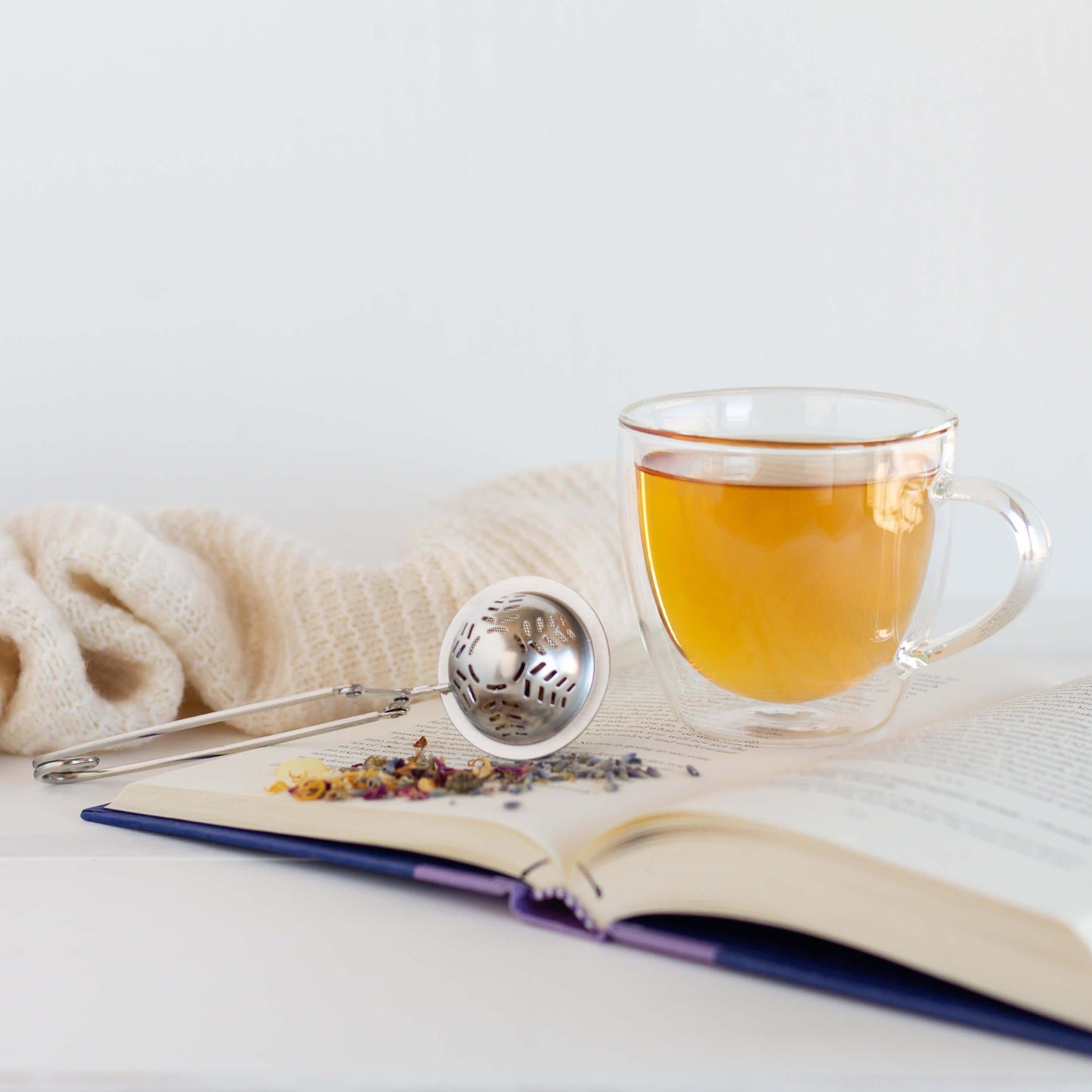 Lavender Lullaby Organic Herbal Tea shown as brewed tea in a glass mug displayed on an open book with a teaball, loose lavender blossoms, and a white knitted blanket in the background