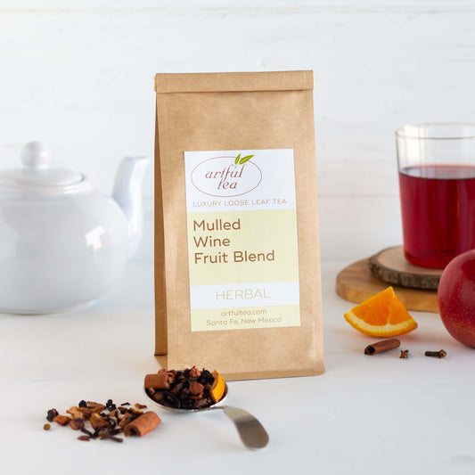 Mulled Wine Fruit Blend Herbal Tea shown packaged in a brown Kraft bag with ingredients nearby, a cup of brewed tea on a wooden coaster, and a white teapot.