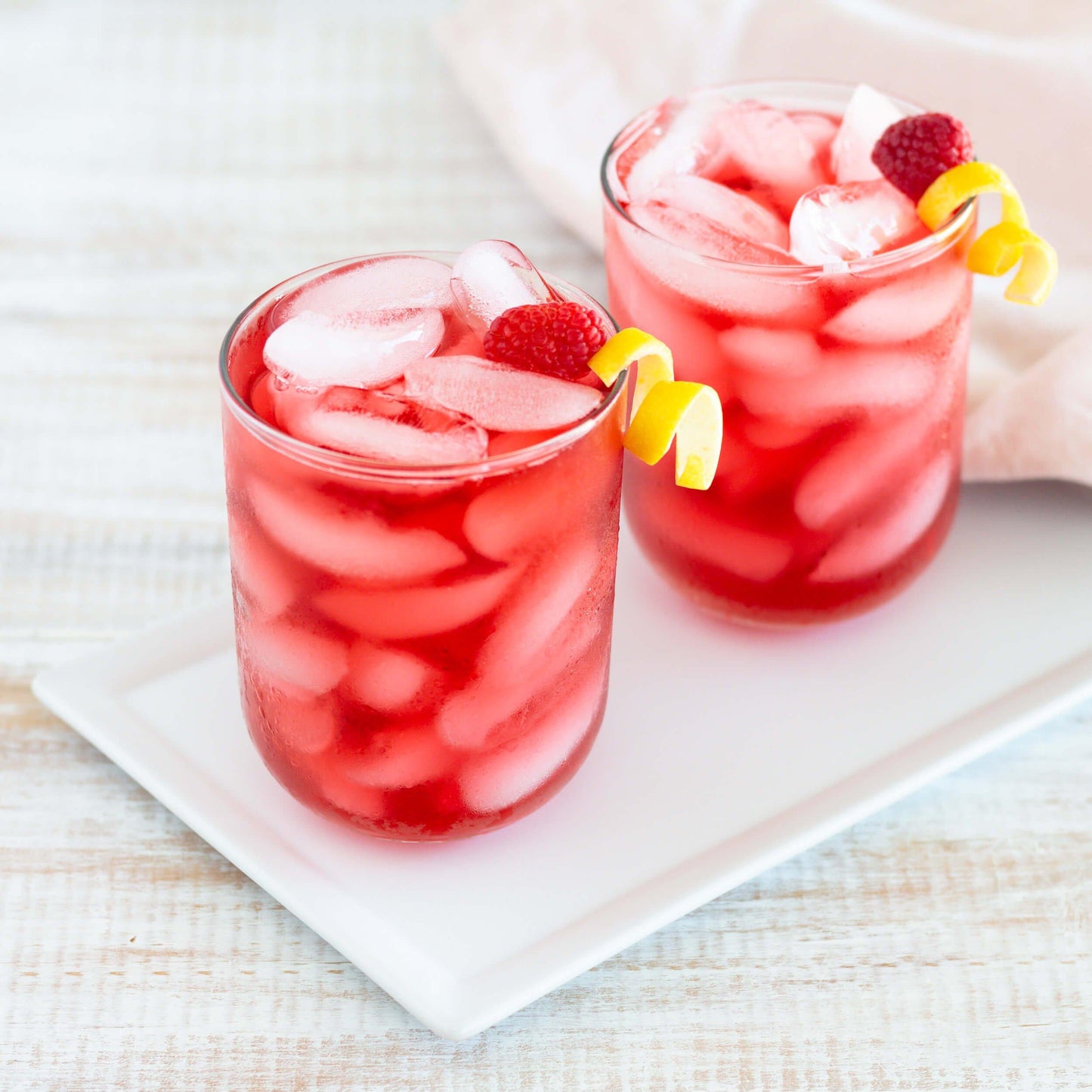 Raspberry Lemonade Organic Herbal Tea shown as iced tea in two short glasses each with a twist of lemon and a single raspberry garnish. The glasses are displayed on a rectangular white tray with a white cloth in the background