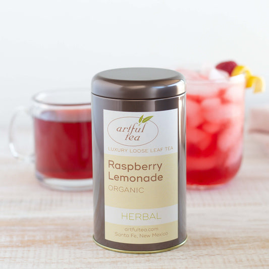 Raspberry Lemonade Organic Herbal Tea shown packaged in a brown tin, with a glass mug of brewed tea and a short glass of iced tea in the background