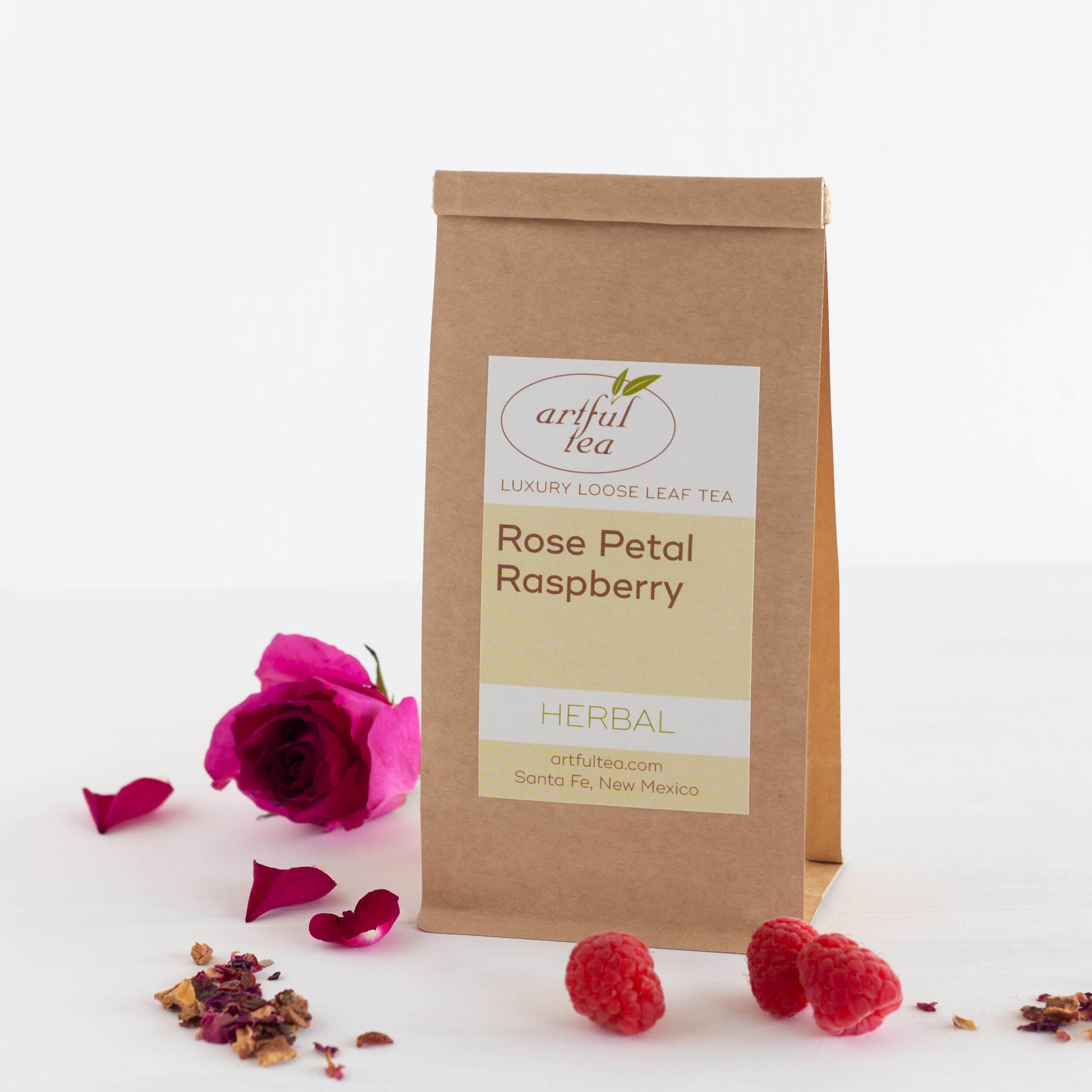 Rose Petal Raspberry Herbal Tea shown packaged in a brown Kraft bag surrounded by raspberries and pink roses.