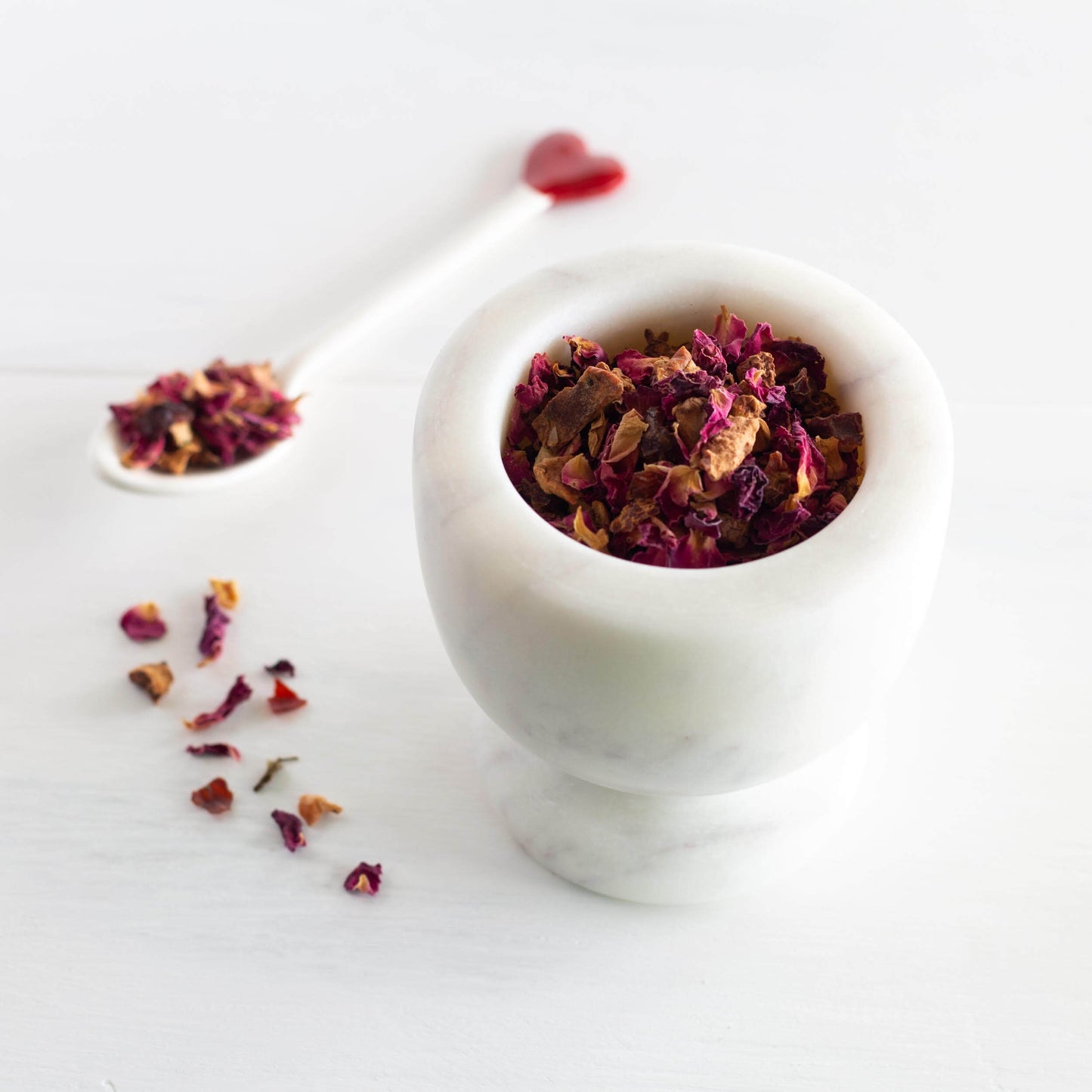 Rose Petal Raspberry Herbal Tea shown as a loose leaf tea inside of a white marble dish with a white spoon of loose tea nearby.