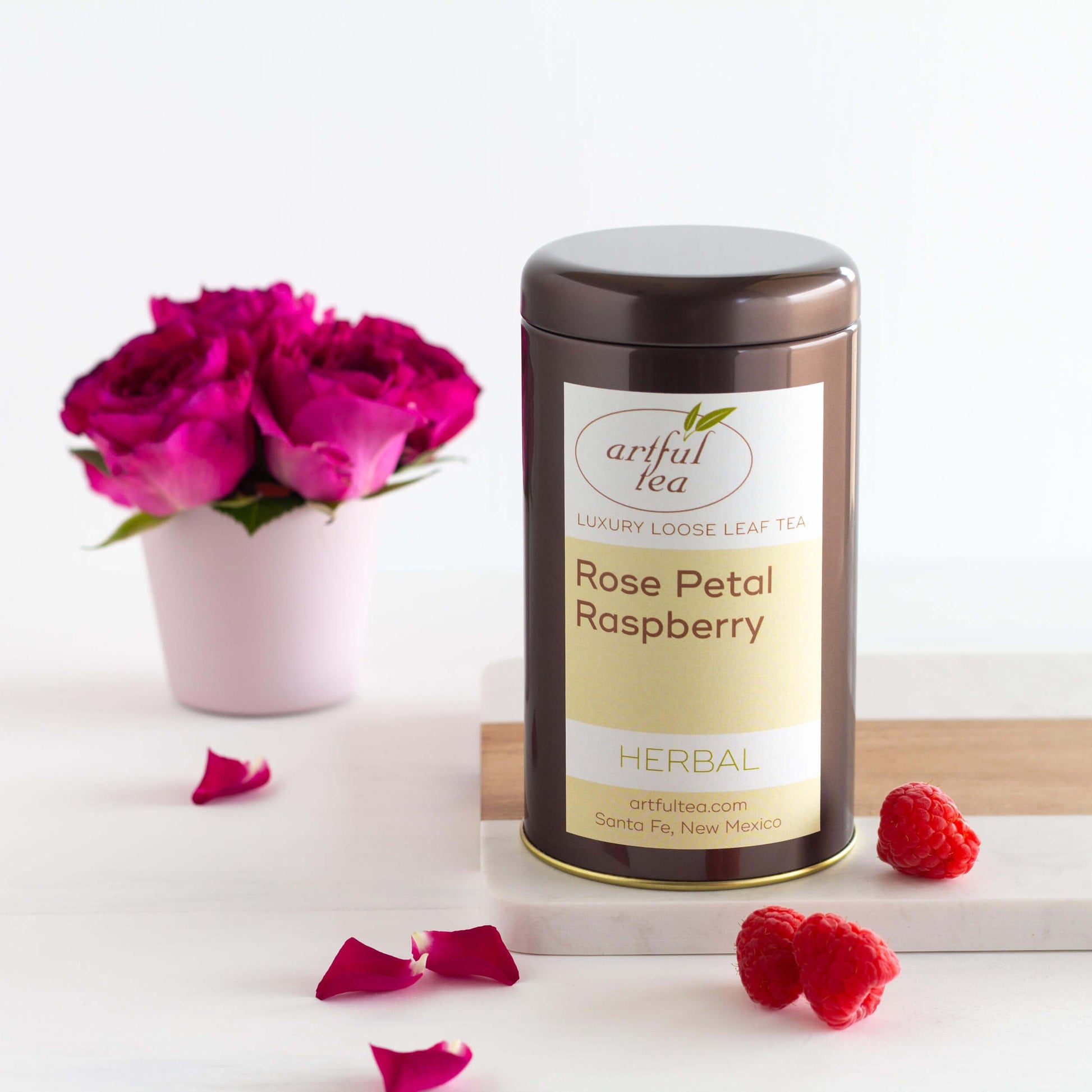 Rose Petal Raspberry shown packaged in a brown tin next to raspberries and pink roses.