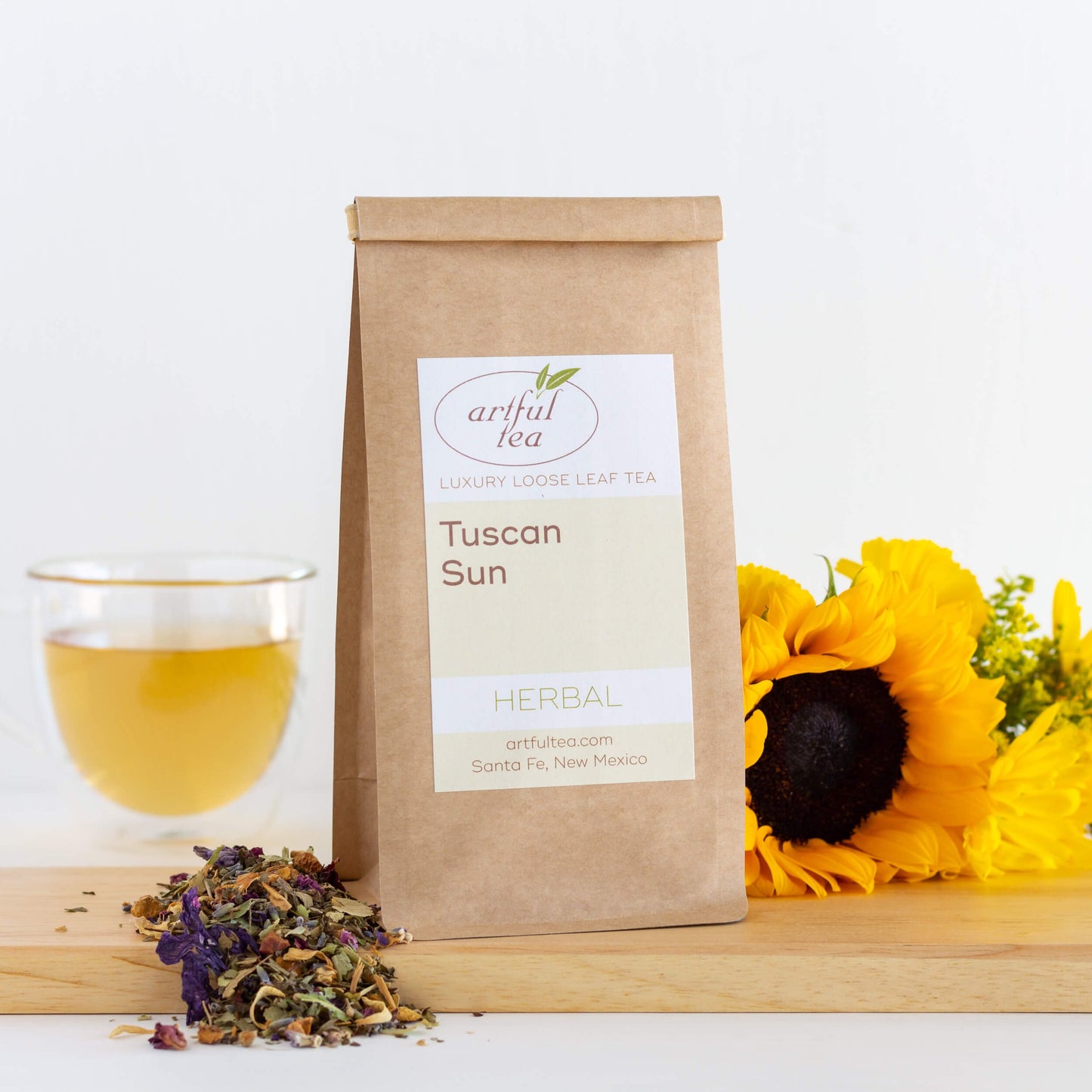 Tuscan Sun Herbal Tea shown packaged in a brown Kraft bag next to a pile of loose leaf tea and yellow flowers on a wooden plank. In the background is a clear cup of brewed tea.