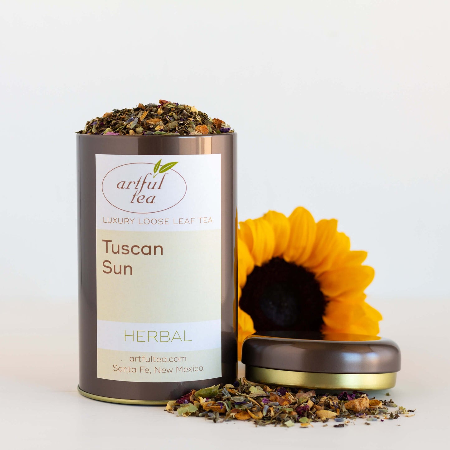 Tuscan Sun Herbal Tea shown packaged in a brown tin with the lid off and a pile of loose tea leaves next to it. Behind everythingis a large sunflower.