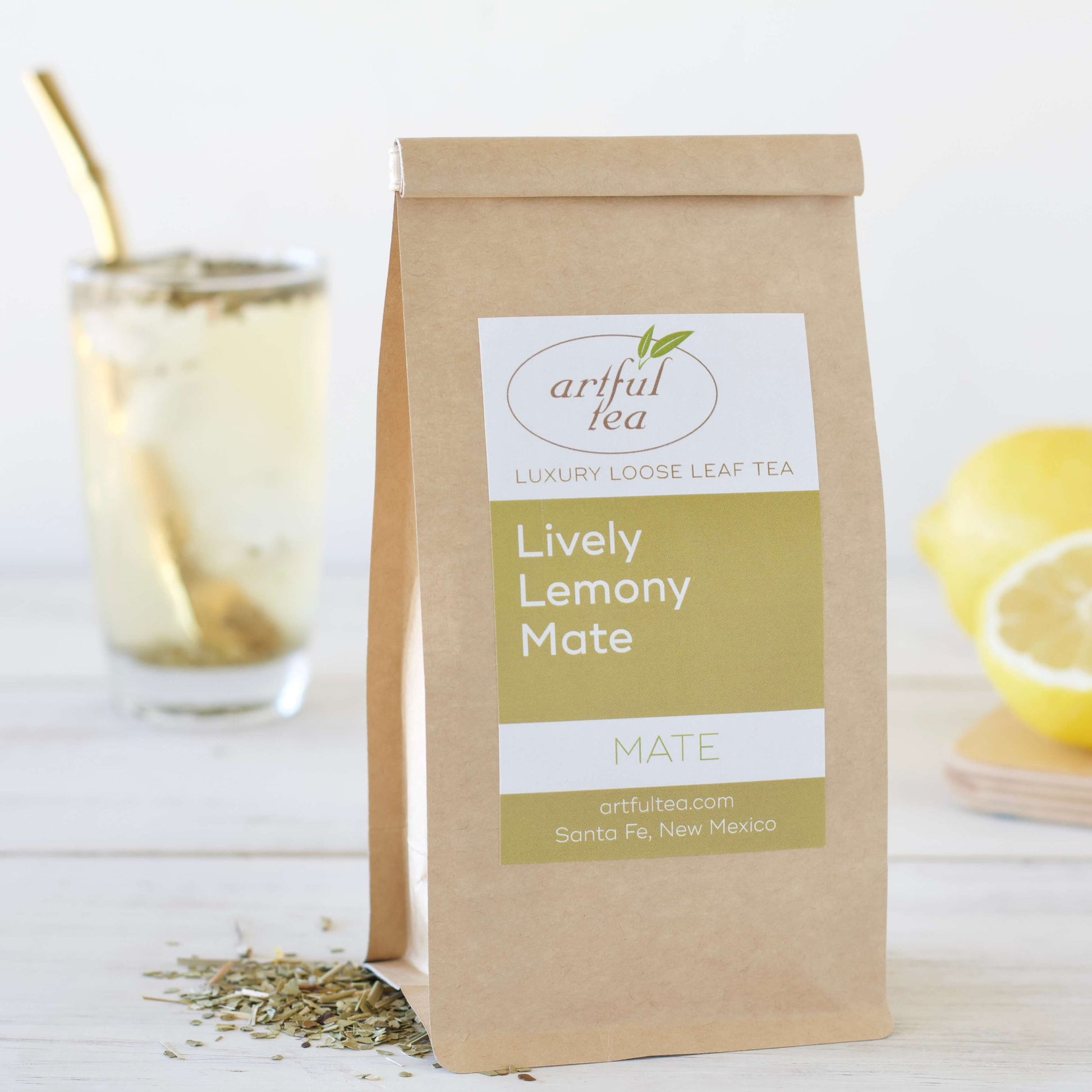 Lively Lemony Mate Tea shown packaged in a brown Kraft bag with loose leaf tea next to it. In the background is a clear glass of brewed tea with a bombilla and lemons.