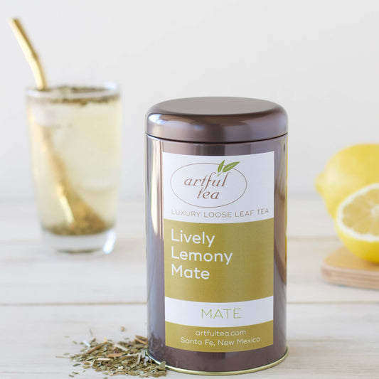 Lively Lemony Mate Tea shown packaged in a brown tin with loose leaf tea next to it. In the background is a clear glass of brewed tea with a bombilla and lemons.