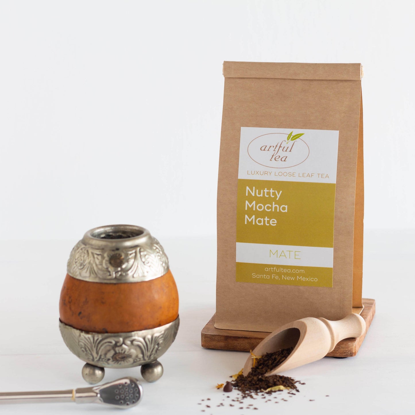Nutty Mocha Mate Tea shown packaged in a brown Kraft bag on top of a wooden coaster with a wooden scoop next to it that has loose leaf tea spilling out. Beside it is a mate gourd and bombilla.