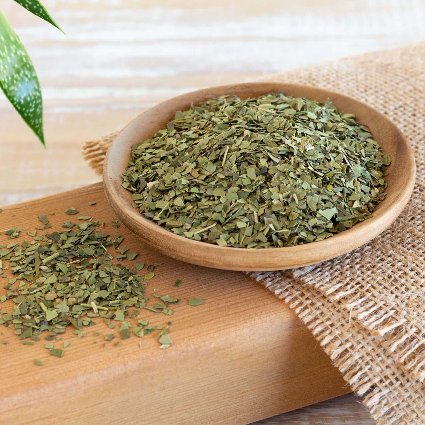 Yerba Mate Tea shown as loose leaf tea in a wooden bowl on top of a burlap which is all on top of a wooden plank, with more loose tea on the plank.