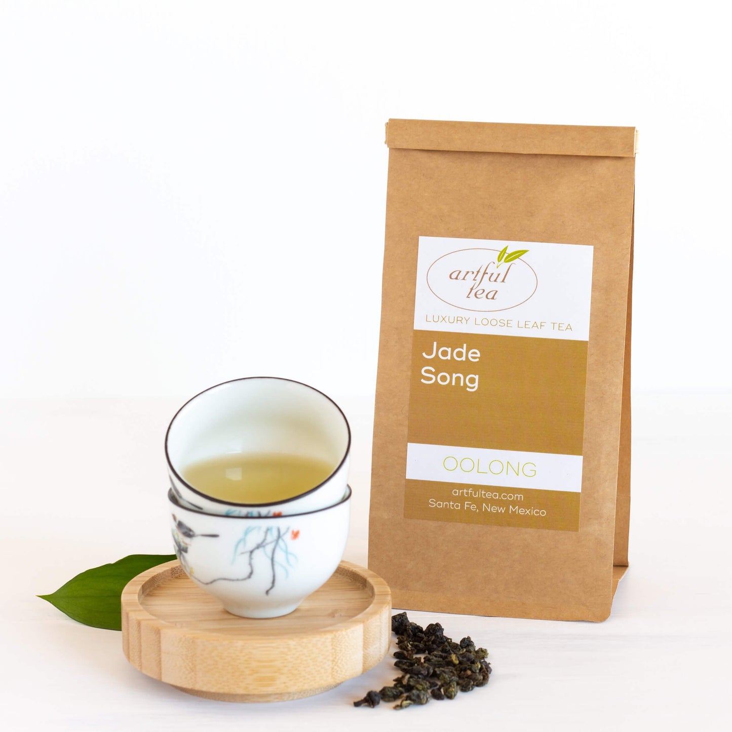 Jade Song Oolong Tea shown packaged in a brown Kraft bag next to a white cup of brewed tea on top of a brown coaster, with loose leaf tea next to it.