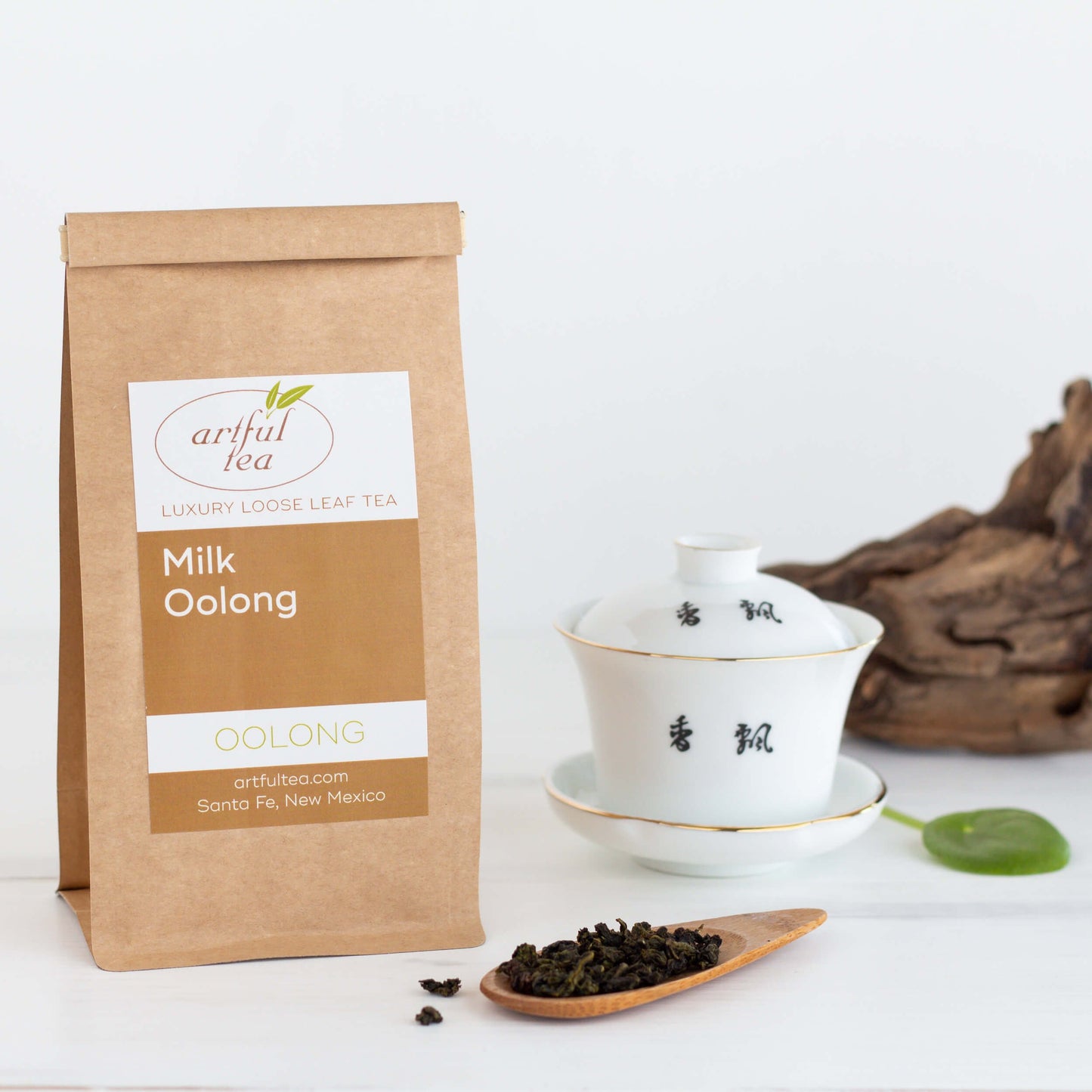 Milk Oolong Tea shown packaged in a brown Kraft bag beside a white Taiwan cup and a wooden spoon of loose leaf tea.