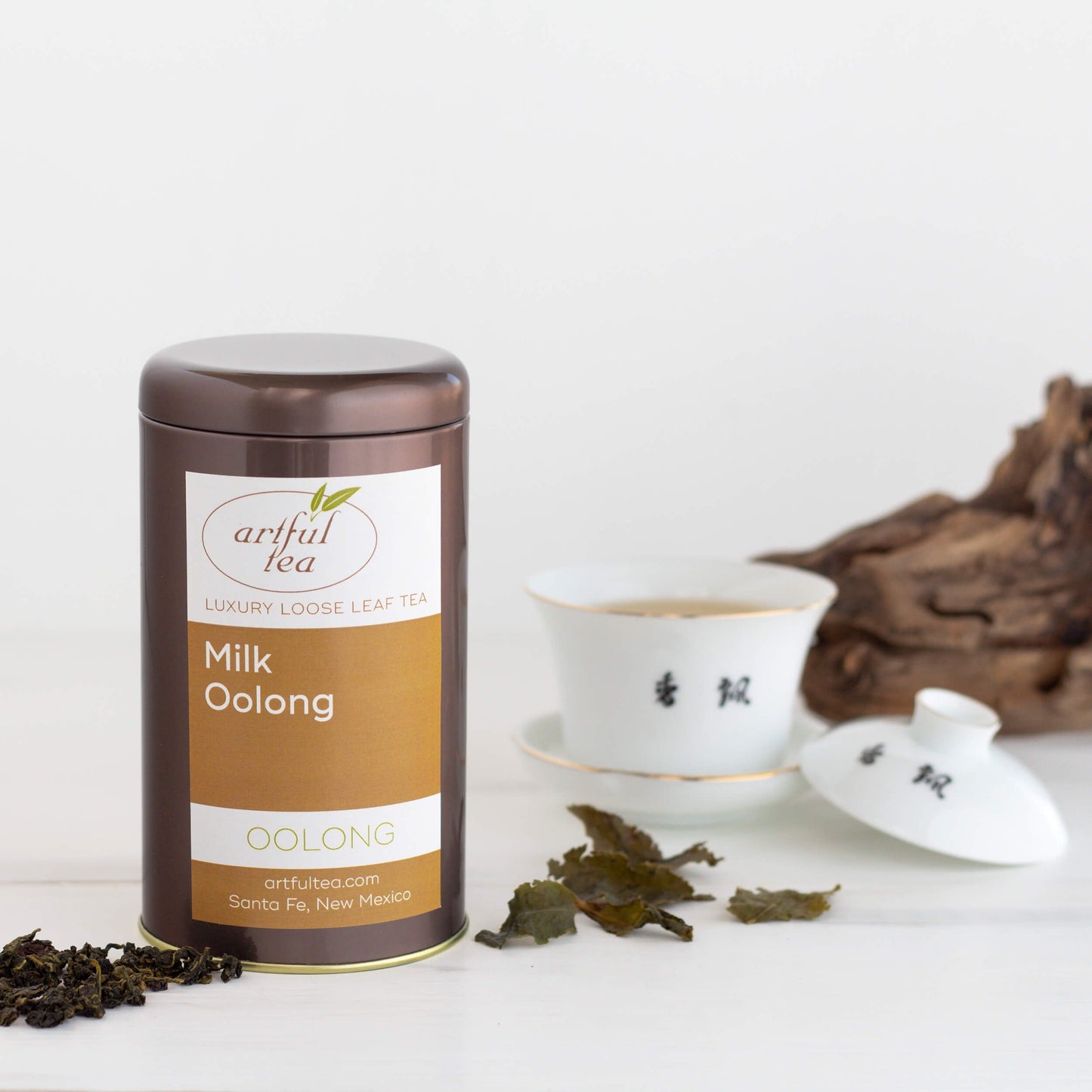 Milk Oolong Tea shown packaged in a brown tin with loose leaf tea around it. In the background is a white Taiwan cup with the lid off and a piece of wood.