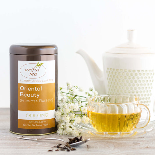 Oriental Beauty Oolong Tea shown packaged in a brown tin beside a clear mug of brewed tea, a large white teapot, and a bundle of baby's breath flowers.