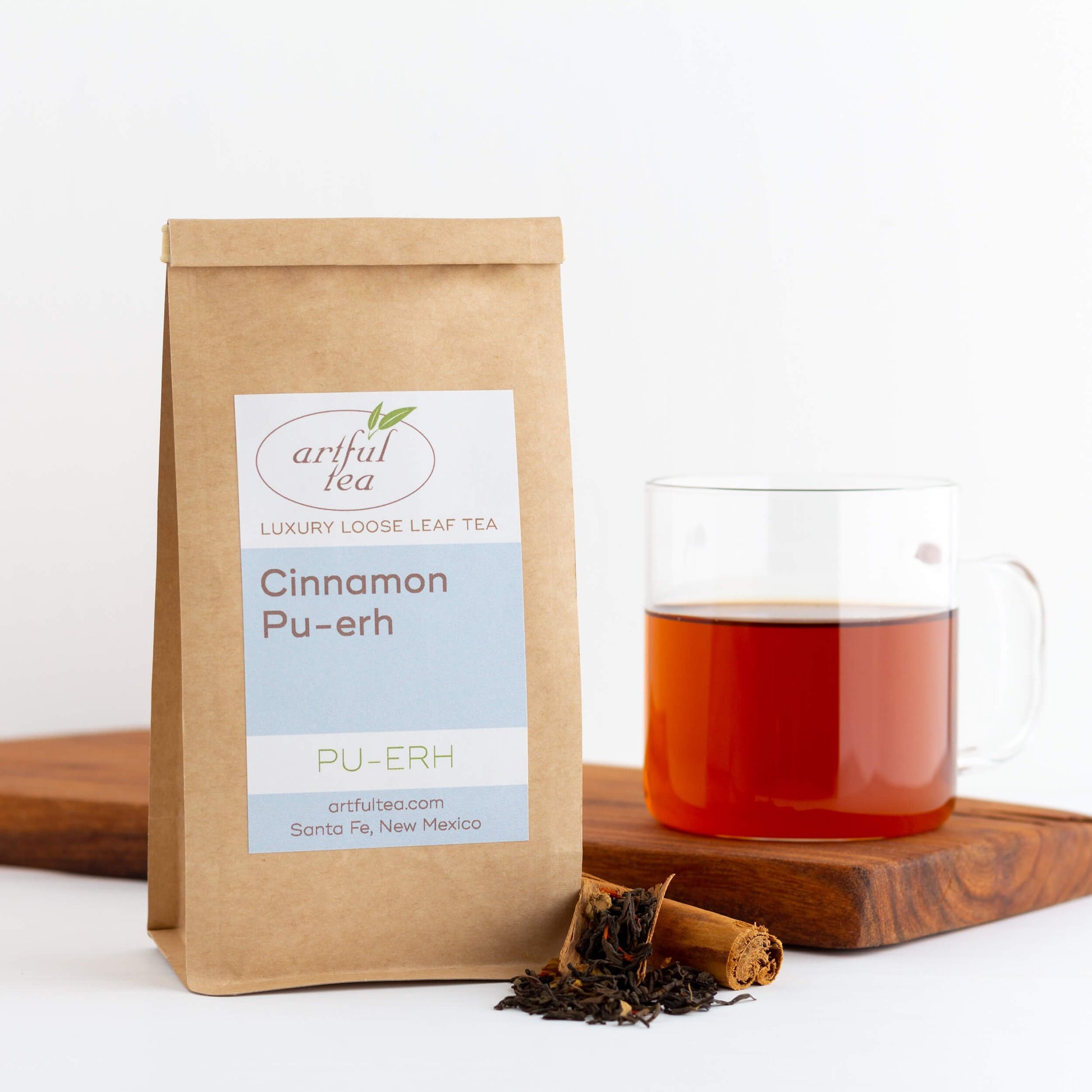 Cinnamon Pu-erh Tea shown packaged in a brown Kraft bag with ingredients next to it. Nearby is a clear mug of brewed tea on top of a wooden plank.