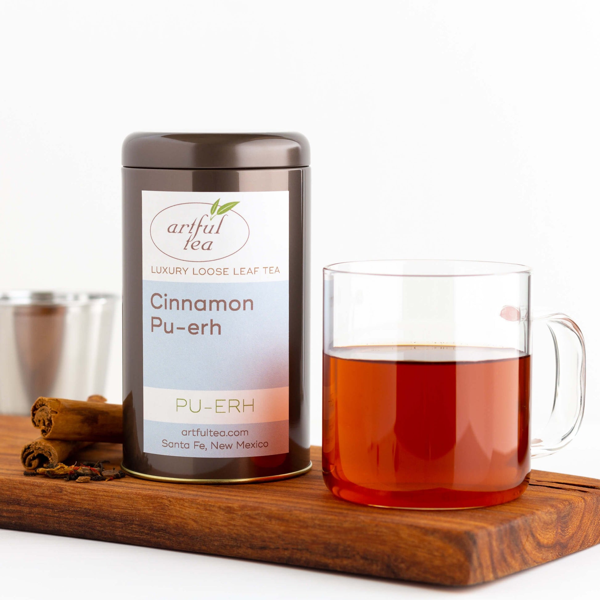 Cinnamon Pu-erh Tea shown packaged in a brown tin next to a clear mug of brewed tea and cinnamon sticks, all on top of a wooden plank.