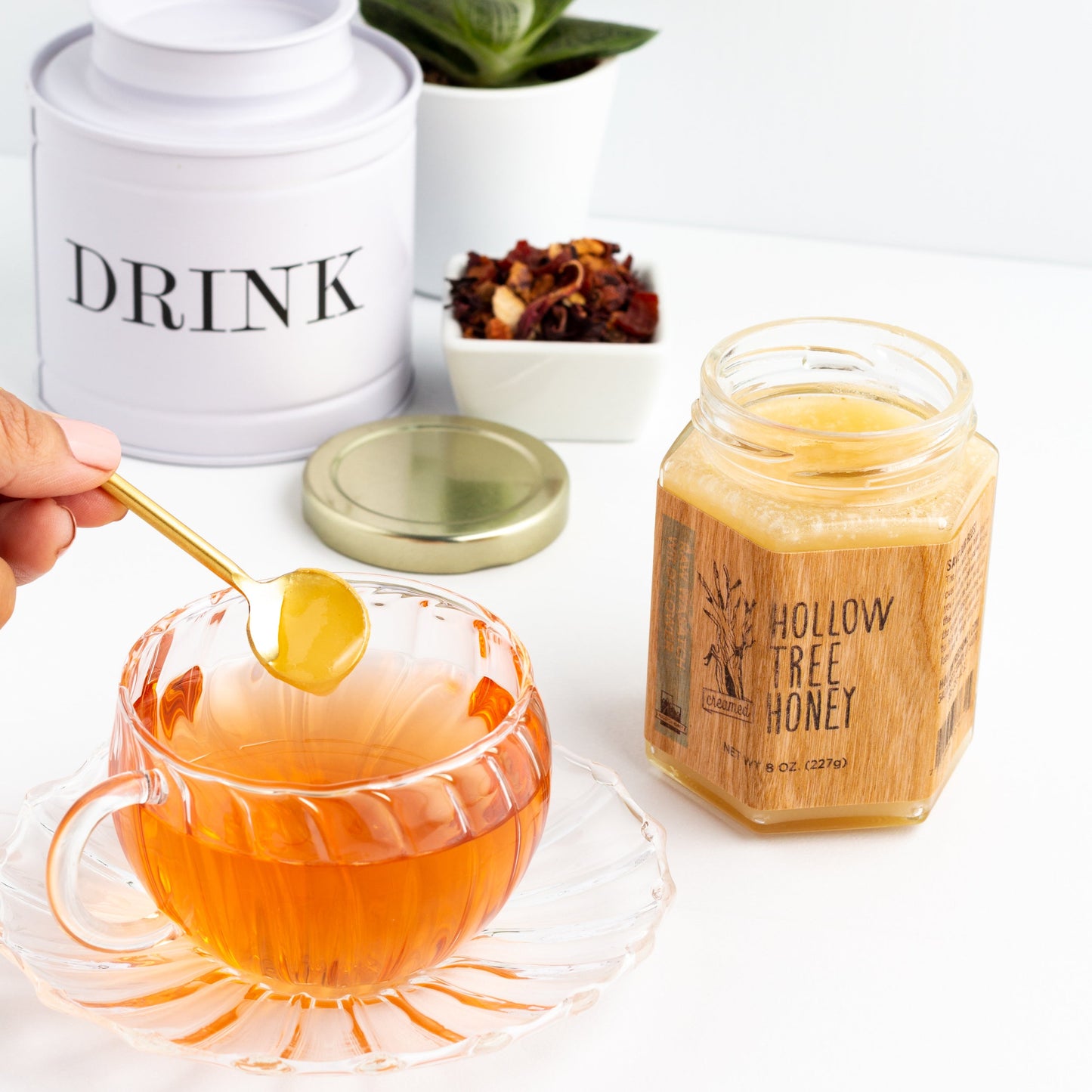 Hollow Tree Honey creamed honey shown in an open glass jar next to a glass teacup with a hand about to stir a small spoonful of honey into the tea