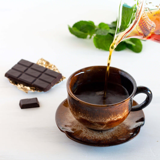 Chocolate Mint Rooibos Herbal Tea being poured into a brown teacup on a saucer, with a bar of chocolate nearby