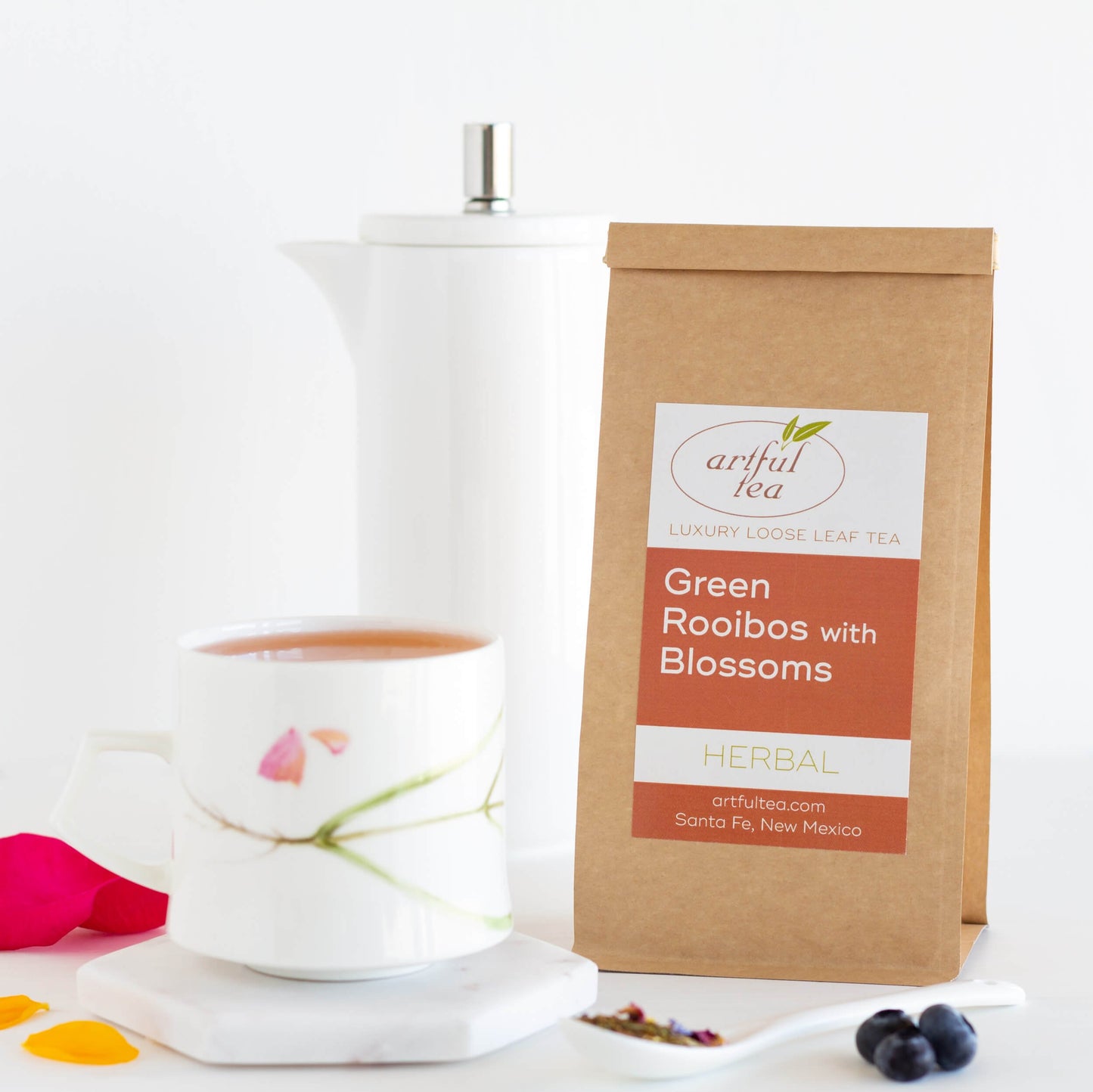 Green Rooibos with Blossoms Herbal Tea shown packaged in a brown Kraft bag beside a white mug full of brewed tea on a white coaster. In the background is a white tall pitcher, in the foreground is a white spoon of loose leaf tea and blueberries.