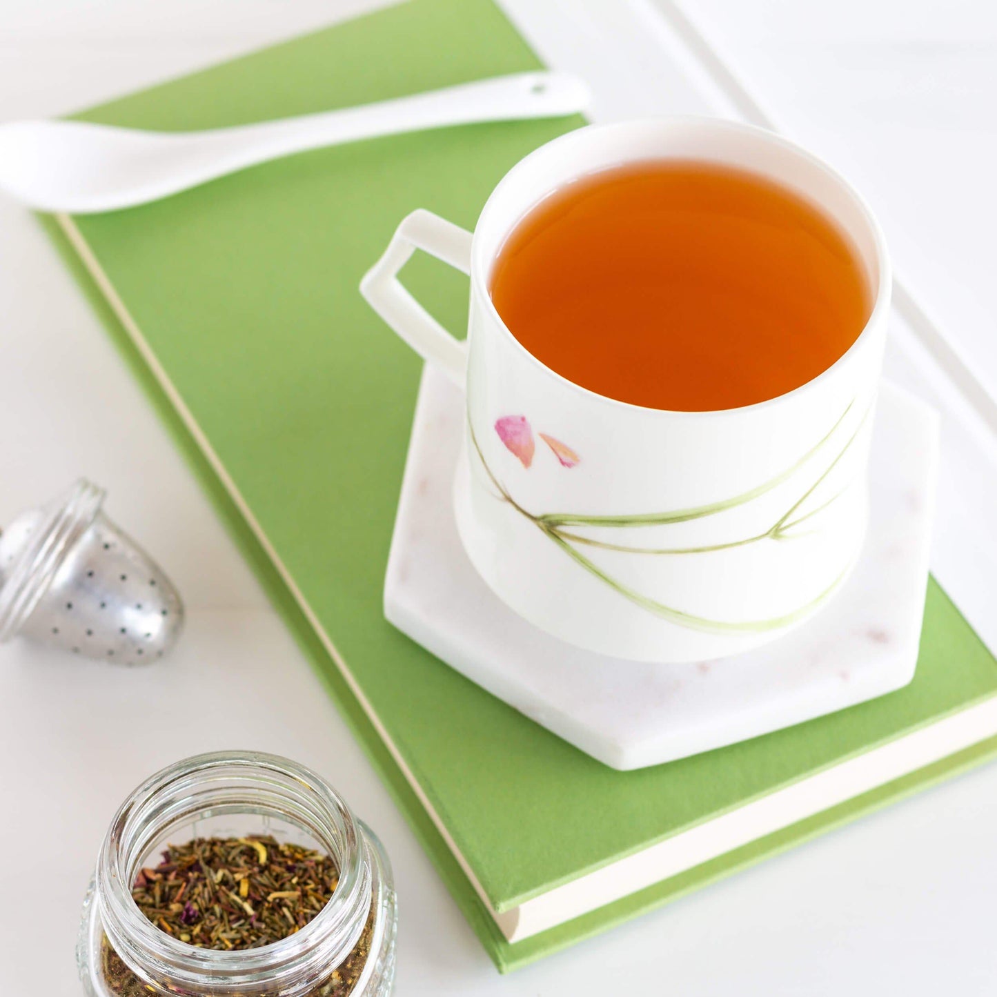 Green Rooibos with Blossoms Herbal Tea shown brewed in a white mug with a white spoon on top of a green book.