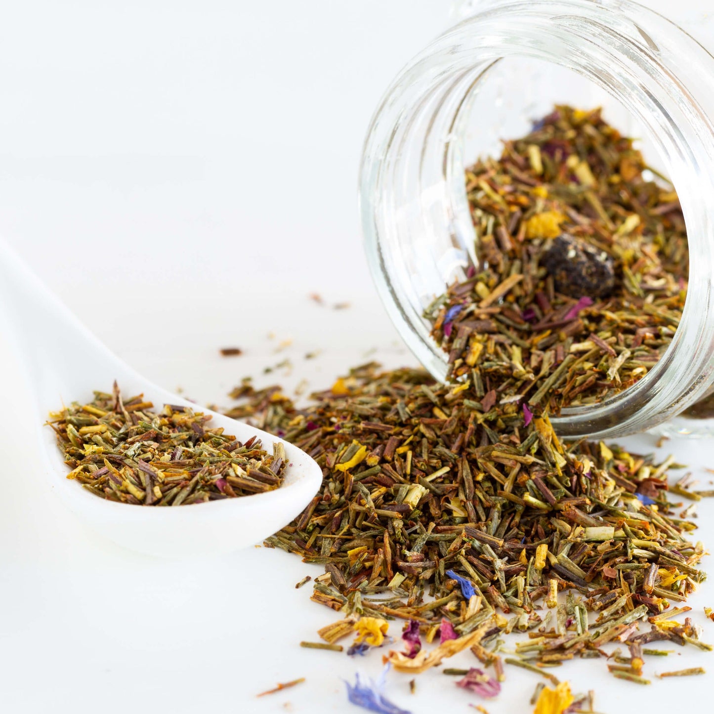 Green Rooibos with Blossoms Herbal Tea shown as loose leaf tea spilling out of a clear jar next to a white spoon of loose tea.