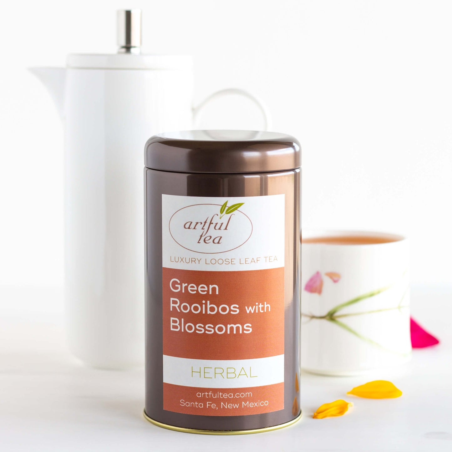 Green Rooibos with Blossoms Herbal Tea shown packaged in a brown tin. Behind it is a white cup of brewed tea next to a tall white pitcher.