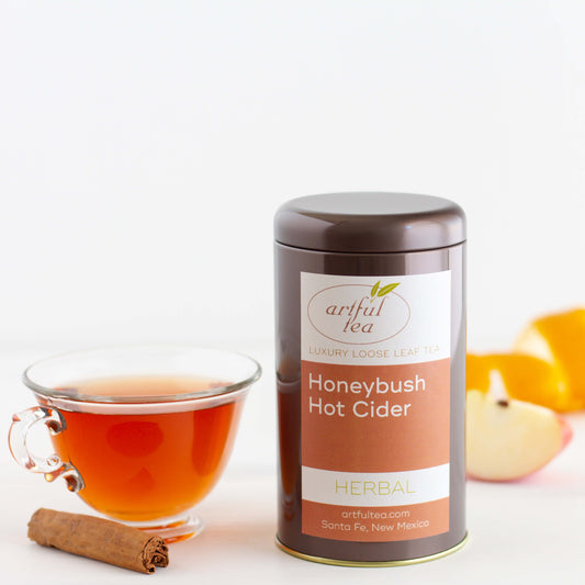 Honeybush Hot Cider Herbal Tea shown packaged in a brown tin next to a clear mug of brewed tea surrounded by ingredients.