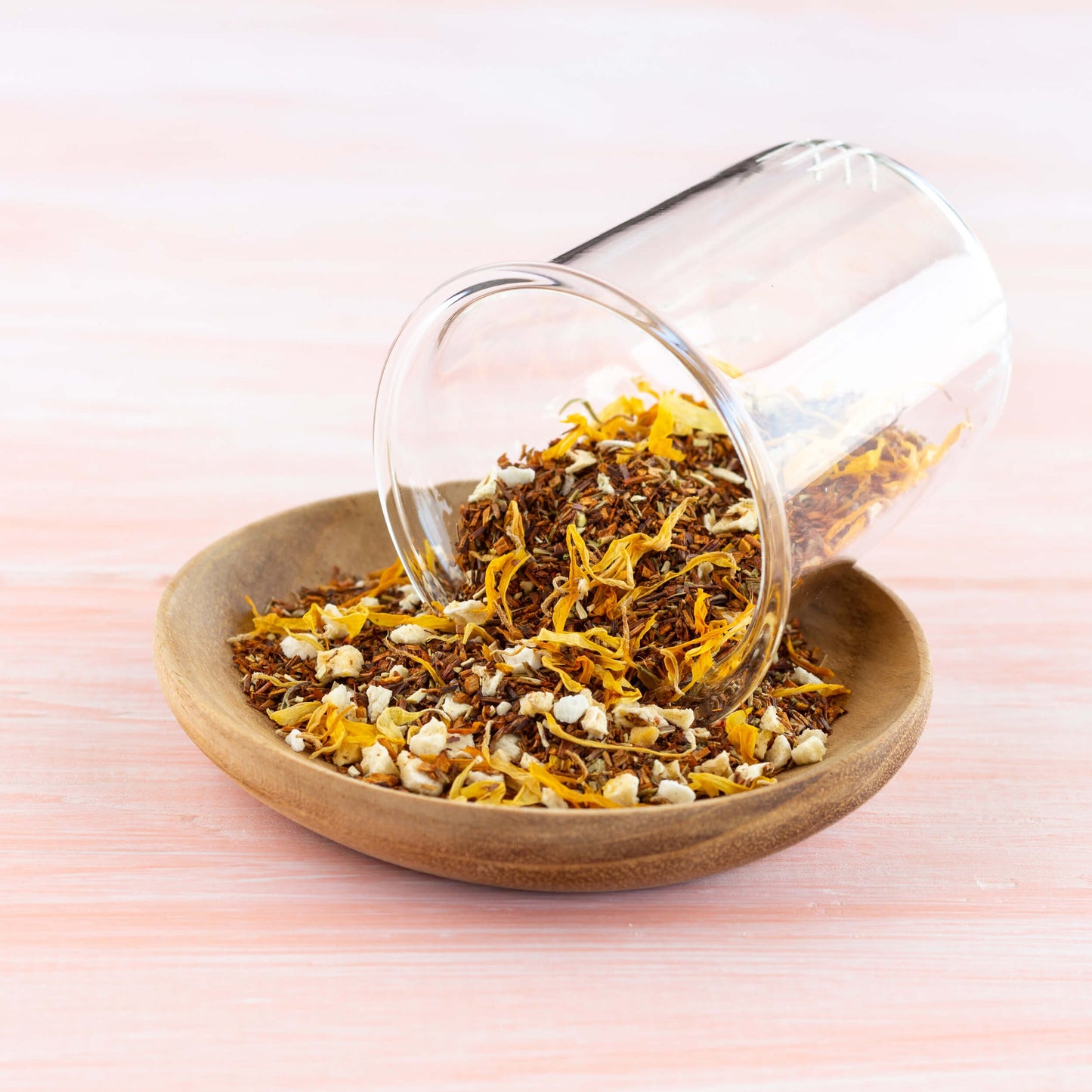 Peach Rooibos Herbal Tea shown as loose leaf tea spilling out of clear cup and into a wooden bowl.