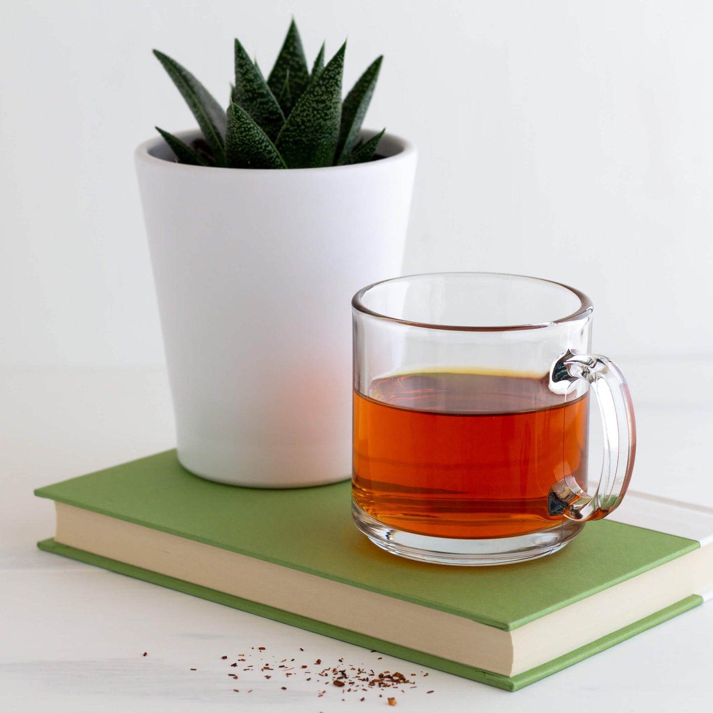 Rooibos Herbal Tea shown as brewed tea in a clear mug next to a plant in a white pot on top of a green book.