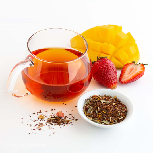 Strawberry Mango Rooibos Herbal Tea shown brewed in a clear mug next to a mango, strawberries, and a small bowl of loose leaf tea.