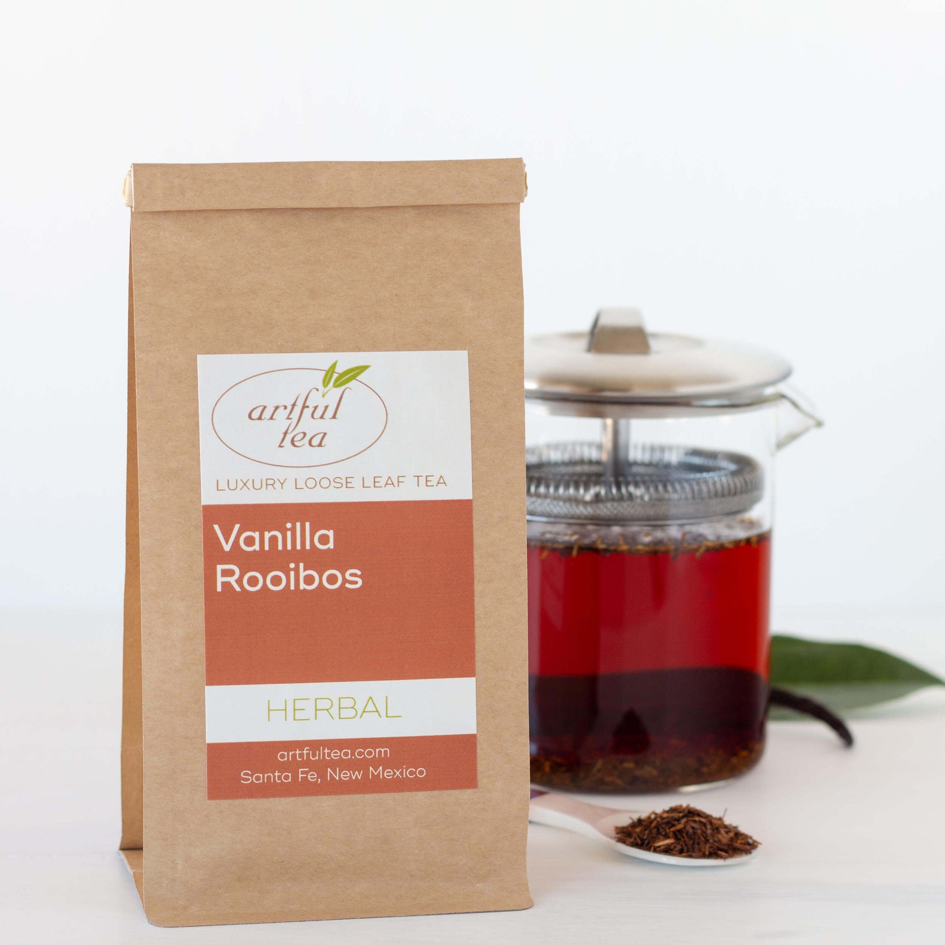 Vanilla Rooibos Herbal Tea shown packaged in a brown Kraft bag beside a small clear French press full of vanilla rooibos and a white scoop of loose leaf tea.