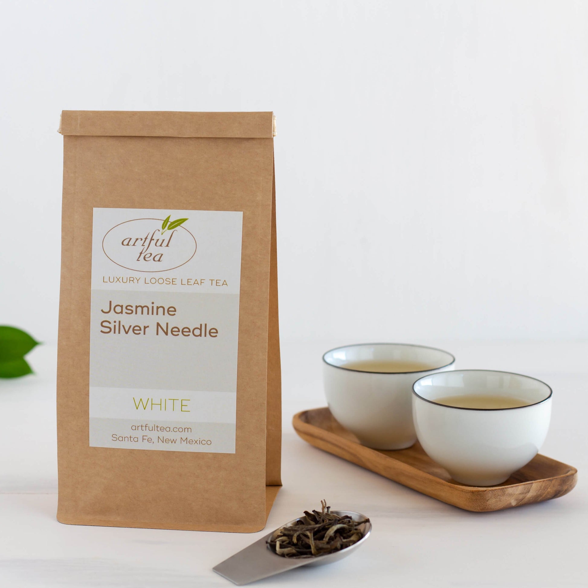 Jasmine Silver Needle White Tea shown packaged in a brown Kraft bag next to a silver scoop of loose tea leaves. Also next to it are two white cups of brewed tea on a wooden tray.