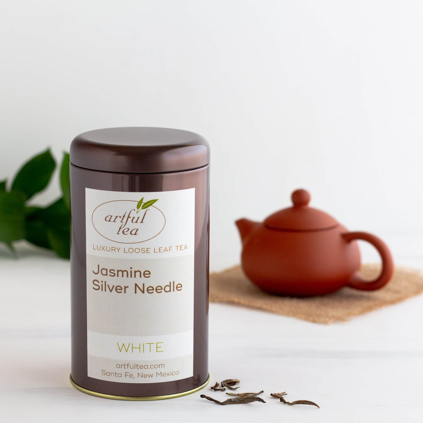 Jasmine Silver Needle White Tea shown packaged in a brown tin next to a few loose tea leaves. In the background is a brown tea pot on a burlap square.