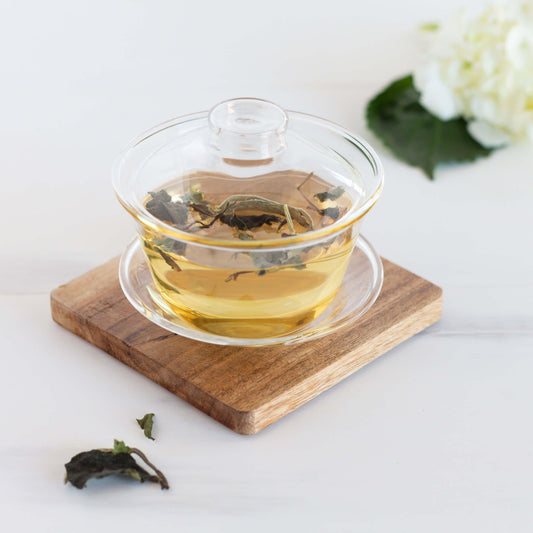 White Peony Organic White Tea shown as brewed tea in a glass gaiwan cup with a lid, displayed on a wooden coaster